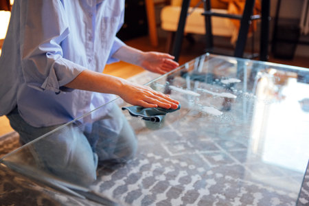 A young woman kneels to wipe a glass table with a cloth. The girl cleans the coffee table using a...
