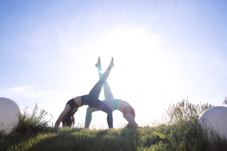 Two female yogis are performing a bridge pose in a tranquil outdoor environment, surrounded by grass...