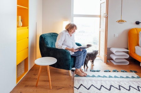 An elderly woman is sitting in an armchair and reading a novel. A middle-aged lady is smiling and holding a book. The pensioner is resting at home. Stylish, cozy interior of house and a beloved petの写真素材