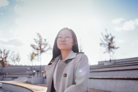 A young Asian woman stands outdoors in a light coat and looks confidently ahead. A girl poses on the...