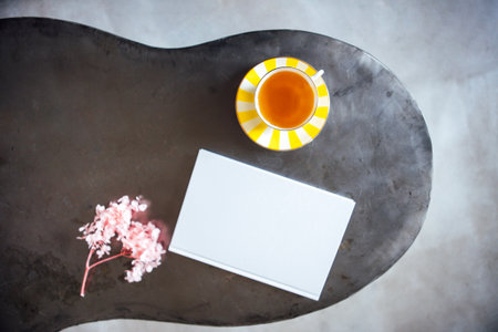 A flat lay scene showcases a white notebook, a yellow-striped tea cup, and pink flowers on a stylish table, evoking a calm and inviting workspace ambianceの写真素材