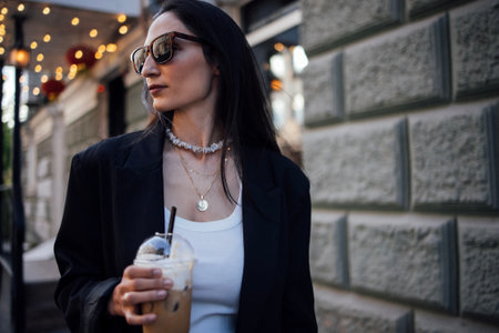 Fashionable woman with dark hair, wearing sunglasses and a black blazer, enjoys a beverage while strolling in a vibrant urban environment, reflecting contemporary styleの写真素材