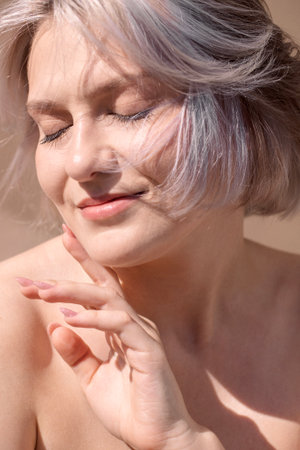 Young woman with silver hair is softly touching her face, highlighting her natural beauty and glowing skin, illuminated by warm sunlight in a calm settingの写真素材