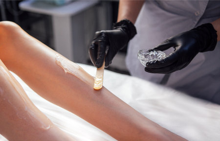 Esthetician is performing hair removal on a client's leg using wax and spatula, highlighting a...