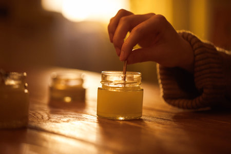 Woman hand is stirring wax in a glass jar on a wooden table, with additional jars nearby, creating a cozy ambiance for candle craftingの写真素材