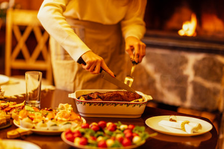 Close-up of a woman cutting a baked duck for a festive dinner. A table with delicious Christmas dishes. The lady cuts off pieces of meat. A fireplace and a cozy home interior in the backgroundの写真素材