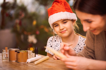 A little girl in a red Christmas hat sits at a table and looks at a sister who decorates candles. A child and a teenager make Christmas gifts. Family creativity and preparation for the holidayの写真素材