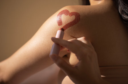 Female figure is using lipstick to draw heart shape on her shoulder, highlighting personal artistry and beauty in a cozy, softly lit environmentの写真素材