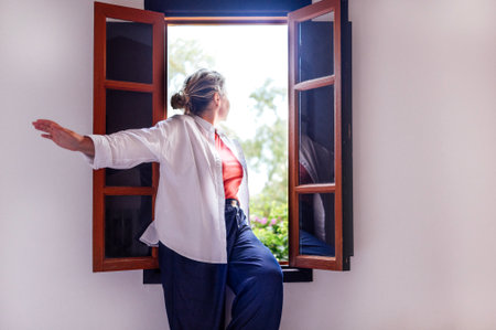 Female figure with light hair leans out of window, embracing fresh air and sunlight, surrounded by greenery, evoking a peaceful and tranquil momentの写真素材