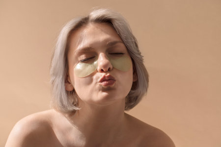 Female with silver hair is applying green eye patches, highlighting a skincare routine against a soft beige background, promoting beauty and self-careの写真素材