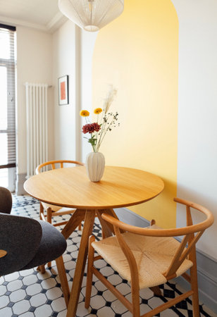 A stylish dining room with a round wooden table with a vase and comfortable chairs made of natural materials. Contrasting interior with white walls and bright yellow details. Modern kitchen designの写真素材