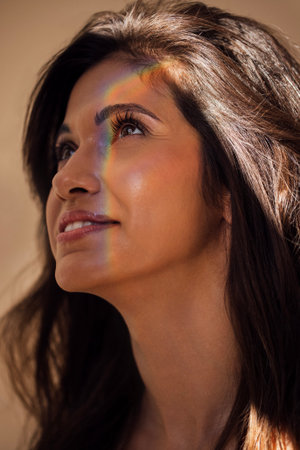 Female with dark hair looks up, bathed in warm light, with a rainbow reflection on her cheek,...