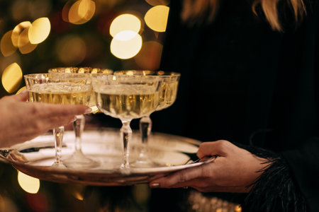 Female server presents champagne glasses on a tray, with sparkling bokeh lights in the background, enhancing the festive mood for celebrationsの写真素材