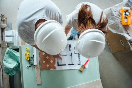 Construction professionals, a man and a woman, are examining detailed blueprints on a table, surrounded by tools and materials, emphasizing collaboration and project planningの写真素材