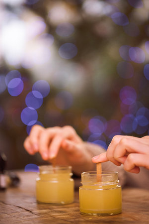 Hands of a woman are mixing yellow candle wax in glass jars on a rustic wooden table, surrounded by soft bokeh lights that enhance the cozy ambianceの写真素材