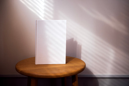 White book positioned on wooden stool, bathed in soft sunlight, with shadows creating a tranquil ambiance, ideal for showcasing design or text with copy spaceの写真素材
