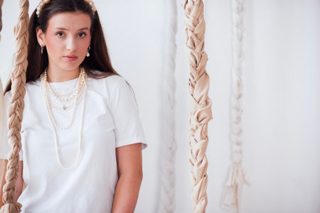 Female model with long hair in a white shirt and pearl necklaces poses elegantly between braided fabric swings, creating a calm and fashionable sceneの写真素材