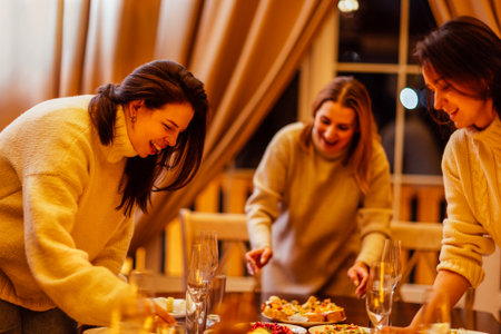 Young women are preparing a Christmas lunch. Friends chat and set the festive table. Girls in sweaters are eating and having fun at a New Year Eve party at homeの写真素材