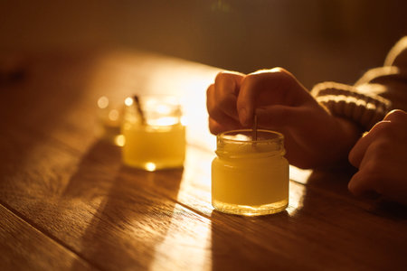 Person's hands are gently stirring a candle jar on a rustic wooden table, with warm sunlight casting soft shadows, enhancing the cozy ambiance of the momentの写真素材