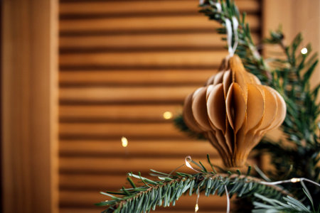 Paper ornament is beautifully suspended from a pine branch, surrounded by warm wooden textures and soft glowing lights, enhancing the festive ambianceの写真素材