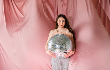Female figure with long dark hair, dressed in shimmering attire, embraces a disco ball in front of a...