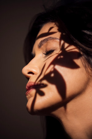 Woman's profile with dark hair, softly lit, features intricate shadows from plant leaves, enhancing the artistic and tranquil ambiance of the sceneの写真素材