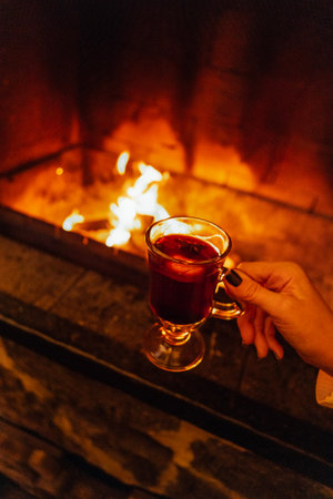 Close-up of a female hand with hot mulled wine. A woman in a sweater holds a delicious fragrant Christmas drink with spices and orange. Fireplace in the backgroundの写真素材