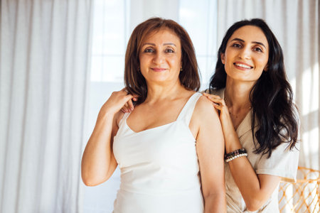 Two women are standing together in a well-lit room, smiling and enjoying each other's company, with soft curtains creating a warm atmosphereの写真素材