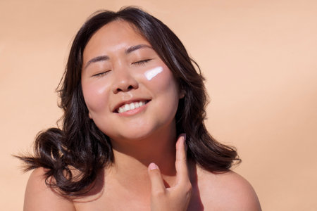 Smiling female applying cream to her face, eyes closed, radiating joy and confidence, highlighting a...
