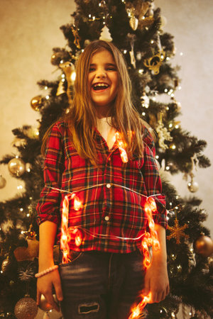 Happy girl with long hair is playfully posing in front of a beautifully decorated Christmas tree, surrounded by festive lights and ornaments, radiating joy and warmthの写真素材