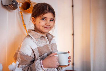 Young girl smiles while holding a warm cup, dressed in a cozy striped sweater, surrounded by charming decor, creating a joyful and inviting ambianceの写真素材