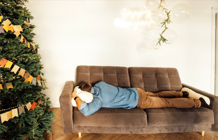 Male individual is lying on a comfortable couch, with a Christmas tree adorned with ornaments nearby, evoking a peaceful holiday spirit and relaxationの写真素材