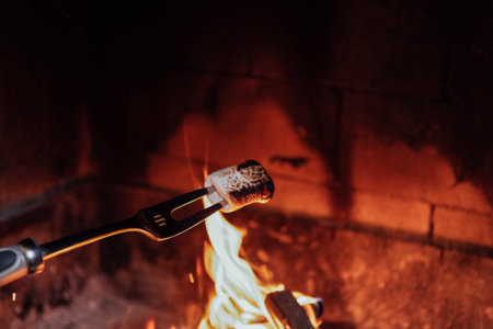 Toasting marshmallow on a fork over a lively campfire, with bright flames and warm embers, creating...