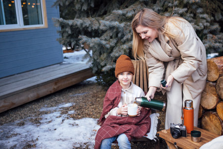 Female adult pours warm drink for child in cozy blanket, seated on bench by logs, with snowy landscape and evergreen trees creating a serene winter atmosphereの写真素材