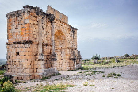 Majestic ancient stone archway ruins of Volubilis, Morocco, are set against a cloudy sky, surrounded by lush grass, highlighting the beauty of historical architecture and its remnantsの写真素材
