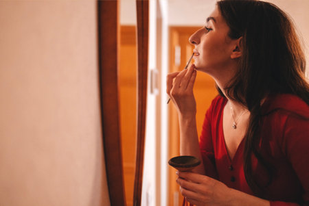 Female applying lipstick, surrounded by warm lighting and elegant decor, highlighting beauty routine and self-care in a serene environmentの写真素材