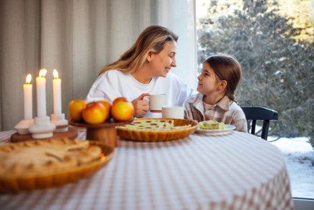 A woman and a girl share a joyful breakfast moment at a table filled with pastries and apples, surrounded by soft natural light and a cozy winter ambianceの写真素材