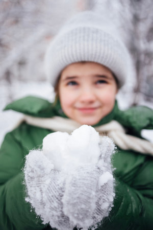 Child with light brown hair in green jacket and gray hat, happily holding snowball in gloved hands, amidst a snowy landscape, evoking winter joy and playfulnessの写真素材