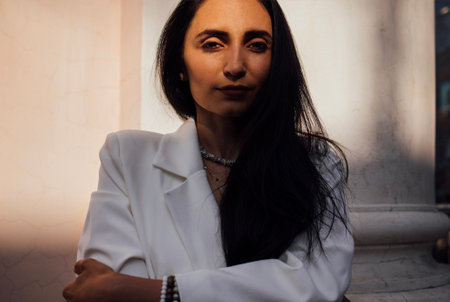Stylish woman with long dark hair, dressed in a white blazer, poses confidently against a textured wall, capturing a moment of elegance and self-assurance in an urban environmentの写真素材