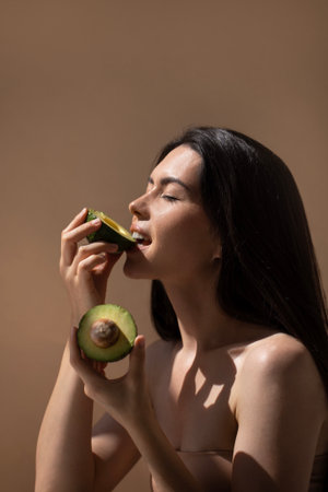 Female model with dark hair is savoring fresh avocado, holding one half while taking a bite from the other, emphasizing health and wellness in a serene settingの写真素材