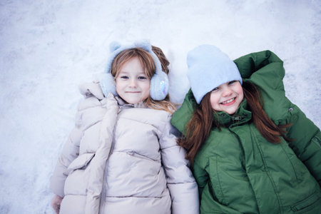 Two children are lying on snow, dressed warmly in winter jackets and earmuffs, enjoying the cold weather and creating joyful moments in a winter wonderlandの写真素材