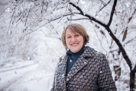 Happy woman with short hair in a fashionable coat stands amidst falling snow, surrounded by trees, embodying the beauty of winter and cheerful spiritの写真素材
