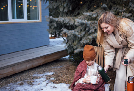 Happy mother  serves warm beverage to child in winter attire, seated on bench outdoors, amidst snow-covered ground and evergreen trees, creating cozy atmosphereの写真素材