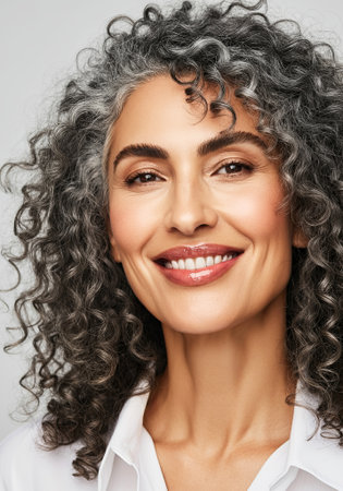 Smiling mature woman with curly gray hair exudes confidence and warmth, highlighting her natural beauty in a professional portrait environment, AI generatedの素材