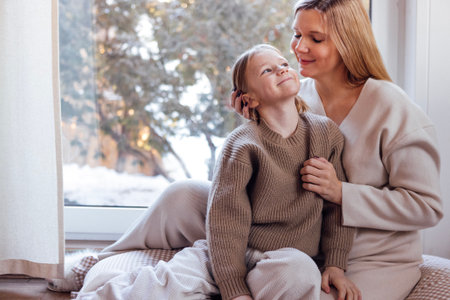 A mother and her child share a tender moment by a window, surrounded by soft textures and warm light, creating a cozy and inviting atmosphere for winterの写真素材