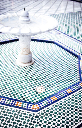Decorative fountain stands in a tiled courtyard, with detailed geometric patterns in green and blue,...