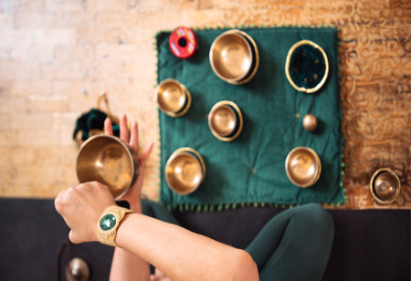 Female musician is playing a Tibetan singing bowl, with multiple bowls arranged on green fabric, evoking tranquility and mindfulness in a peaceful settingの写真素材