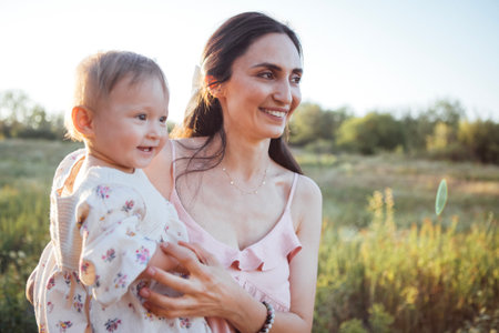 Mother embraces her happy baby girl in a bright field, with lush greenery and soft sunlight creating a warm and loving atmosphere for their special bondの写真素材