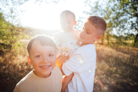 Two boys, one carrying a baby, are smiling in a bright outdoor environment, with sunlight filtering through trees, showcasing a joyful family momentの写真素材