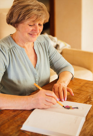 Older female artist is drawing work-life balance wheel sitting on the sofa at home. Self-reflection and life planning in a notebook with colored pen, seated at a rustic wooden table, highlighting her passion for art and creativityの写真素材